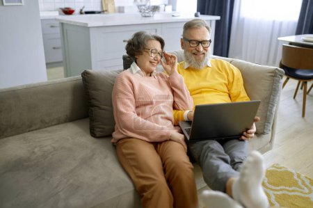 Téléchargez les photos : Couple marié âgé utilisant un ordinateur portable tout en se relaxant à la maison. Les personnes âgées se détendre ensemble lire des nouvelles, faire du shopping en ligne ou regarder des vidéos tout en étant assis sur le canapé à la maison - en image libre de droit