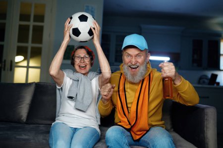 Téléchargez les photos : Joyeux couple de personnes âgées regardant le match de football à la télévision à la maison. mari et femme âgés se sentent ravis et excités hurlant et acclamant favori jeu de football - en image libre de droit
