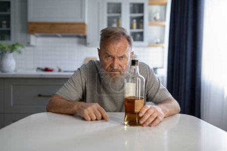 Photo for Portrait of lonely drunk man feeling depressed while sitting at table on home kitchen. Widower with alcohol addiction - Royalty Free Image