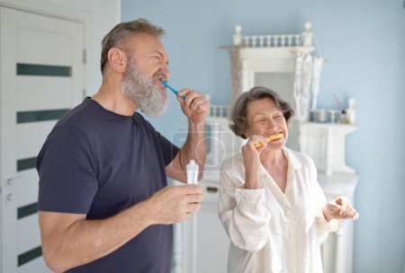 Téléchargez les photos : Couple marié senior brossant les dents ensemble debout à la maison salle de bain. Matin ou soir routine d'hygiène quotidienne de la famille âgée - en image libre de droit