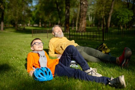 Téléchargez les photos : Grand-père et petit-fils souriants positifs reposent sur la prairie d'herbe verte dans le parc. Mamie soutenant et louant petit-enfant pour de bons patins à roulettes. Happy temps de loisirs en famille le week-end - en image libre de droit
