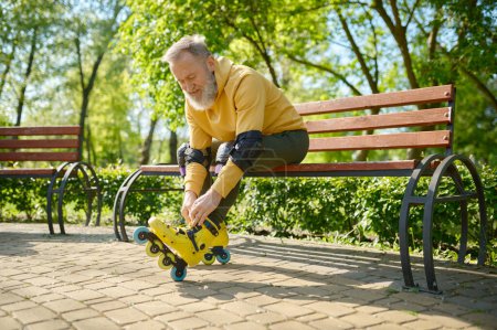 Photo for Grey-haired senior man putting on roller skates while sitting on bench in park. Active recreation time on weekend for elderly people, sport on retirement and healthy lifestyle concept - Royalty Free Image