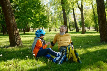 Photo for Happy father and son giving high five each other while rest on green grass meadow in city park after speed racing on rollerskates. Active family weekend - Royalty Free Image