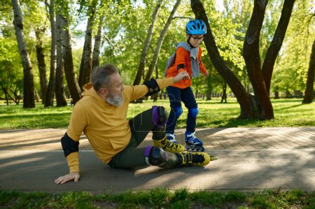Téléchargez les photos : Petit garçon enseignant le patinage à roulettes à son grand-père dans le parc. Grand-père tombant sur la route et petit-fils l'aidant à se lever. Joyeux temps ensemble, enfance heureuse et éducation des enfants - en image libre de droit