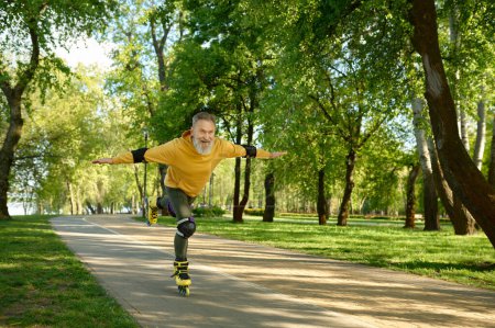 Foto de Alegre anciano emocional haciendo truco mientras monta patines en el parque urbano. Hombre maduro de pelo gris levantando una pierna moviéndose rápido y sintiendo satisfacción. Estilo de vida activo en la jubilación - Imagen libre de derechos