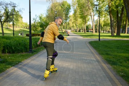 Téléchargez les photos : Homme âgé roulant patins à roulettes regardant en arrière à la caméra. Style de vie sain et concept de roller - en image libre de droit