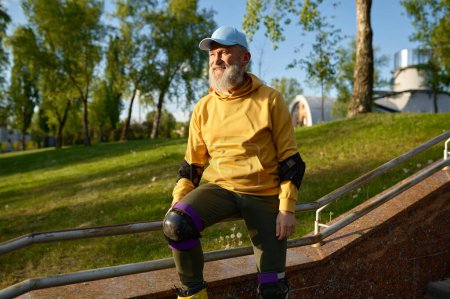 Téléchargez les photos : Heureux homme âgé souriant portant des patins à roulettes repos assis sur une rampe d'escalier dans le parc de la ville. Temps actif à la retraite - en image libre de droit