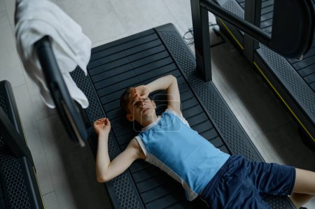 Téléchargez les photos : Fatigué garçon épuisé repos couché sur tapis roulant après un entraînement difficile. Entraînement sportif pour les enfants - en image libre de droit