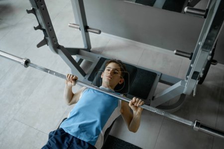 Foto de Un tipo serio y flaco levantando pesadas barras de pesas tumbado en el banco del gimnasio. Concepto de infancia, fitness y deporte - Imagen libre de derechos
