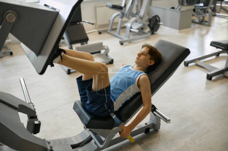 Téléchargez les photos : Préado garçon jambes d'entraînement sur l'équipement sportif à la salle de gym. Jeune athlète masculin poussant la dalle avec les pieds pour être fort et en bonne santé - en image libre de droit