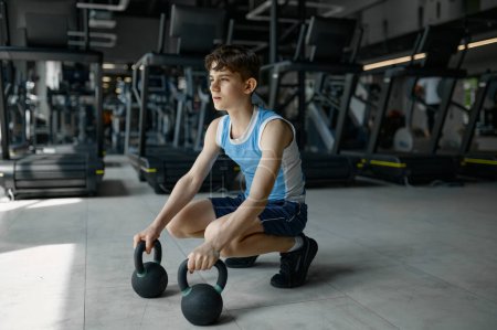 Téléchargez les photos : Adolescent garçon se préparer pour haltérophilie assis avec kettlebell sur sport salle de gym fond - en image libre de droit