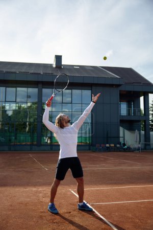 Téléchargez les photos : Jeune joueur de tennis masculin engagé dans l'entraînement sur le terrain de sport de plein air. Homme en tenue de sport frappant balle avec raquette - en image libre de droit