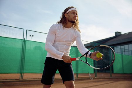 Téléchargez les photos : Joueur de tennis masculin concentré avec raquette et entraînement au ballon sur un terrain extérieur. Préparation pour le service - en image libre de droit