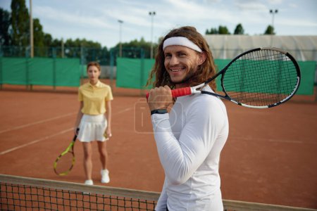 Photo for Friends playing tennis on court. Focus on smiling man player holding racket on shoulder and looking in camera. Weekend sports recreation for young people couple - Royalty Free Image