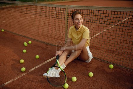Téléchargez les photos : Heureuse joueuse de tennis souriante avec raquette assise sur le terrain parmi les balles éparses. Femme mignonne se détendre après la formation de jeu - en image libre de droit