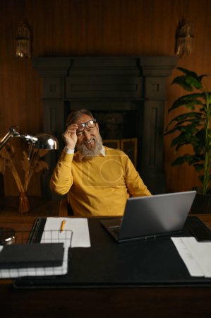 Smiling senior man looking at laptop screen during work or rest at home office in evening. Elderly male entrepreneur holding business conference or watching fun video on computer