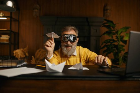 Téléchargez les photos : Drôle réjoui homme mature portant des lunettes de pilote jouant avec l'avion origami tout en étant assis sur le lieu de travail dans le bureau à la maison le soir - en image libre de droit