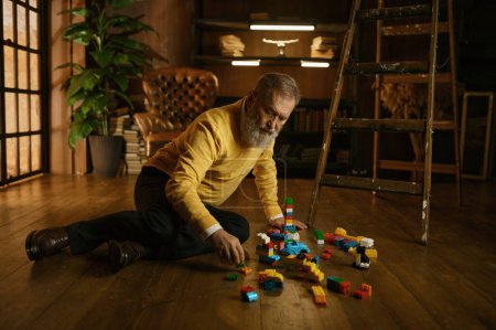 Photo for Senior old man playing toy building blocks while sitting on home floor during cozy evening. Handsome adult businessman on retirement - Royalty Free Image