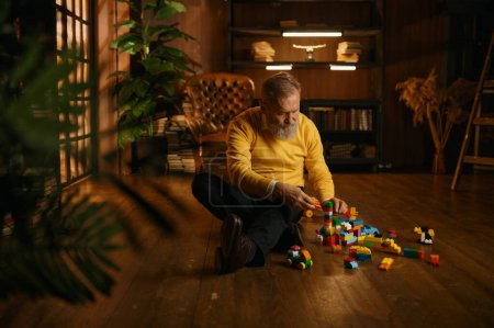 Téléchargez les photos : Vieil homme âgé jouant blocs de construction de jouets tout en étant assis sur le sol de la maison pendant la soirée confortable. Beau homme d'affaires adulte à la retraite - en image libre de droit