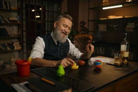 Téléchargez les photos : Drôle élégant homme d'affaires plus âgé fumant du cigare et jouant des jouets à la table de travail dans le bureau à domicile - en image libre de droit