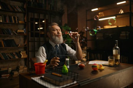 Foto de Divertido hombre de negocios mayor relajado soplando burbujas de jabón mientras está sentado en la mesa de trabajo - Imagen libre de derechos
