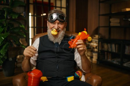 Photo for Portrait of crazy funny senior businessman with toys in action. Mature male wearing elegant costume and pilot goggles feeling fun and joy holding rubber duck in teeth and plane in hand - Royalty Free Image