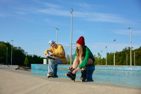 Téléchargez les photos : Préparation à la formation dans le parc. Les jeunes aiment patiner en portant des rouleaux sur les pieds tout en étant assis sur le banc - en image libre de droit