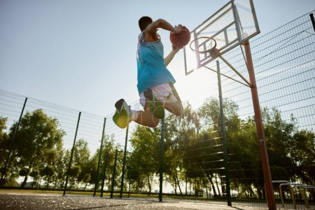 Photo for Teenager player throwing ball into basketball hoop, shot in motion, bottom view. Playing sport game on outdoor court or school playground - Royalty Free Image
