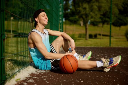 Téléchargez les photos : Satisfait joueur de basket-ball adolescent repos assis sur le terrain de rue. Jeune sportif souriant prendre une pause dans le jeu se sentir bien - en image libre de droit