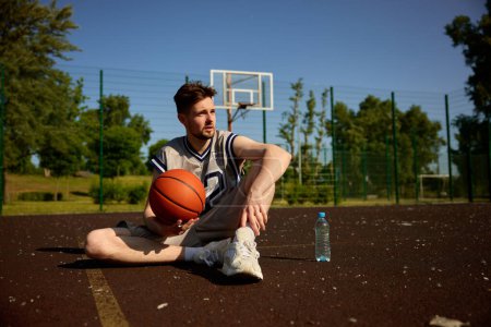 Foto de Joven jugador de baloncesto adulto descansar en la pista urbana. Deportista en uniforme de equipo sosteniendo la pelota relajándose después del juego - Imagen libre de derechos