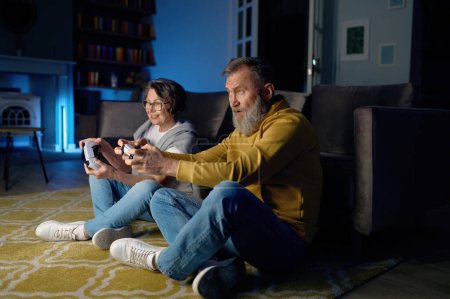 Photo for Senior married couple sitting on floor and playing video game on console. Elderly wife and husband holding gamepad and enjoying challenge each other to win - Royalty Free Image