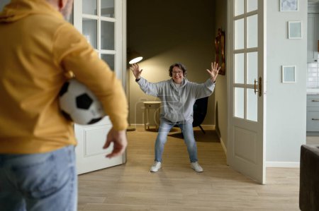 Photo for Joyful family couple of pensioner playing football at home. Selective focus on ball in hand of older man and smiling excited elderly woman ready for pass on blurred background - Royalty Free Image