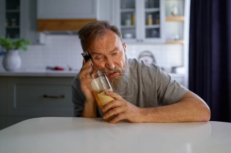 Photo for Portrait of lonely drunk man feeling depressed while sitting at table on home kitchen. Widower with alcohol addiction - Royalty Free Image