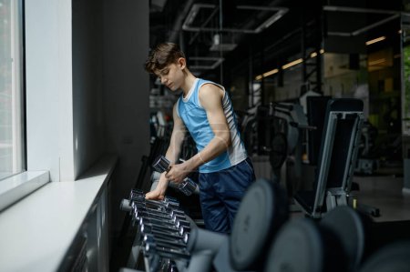 Téléchargez les photos : Adolescent garçon prendre haltère de différents poids de rack préparation pour entraînement séance d'entraînement à la salle de gym - en image libre de droit