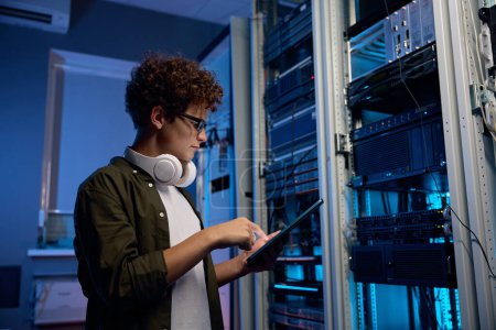 Photo for Serious young man wearing headphones working in data center standing at open server rack cabinet and using mobile tablet to set up system - Royalty Free Image