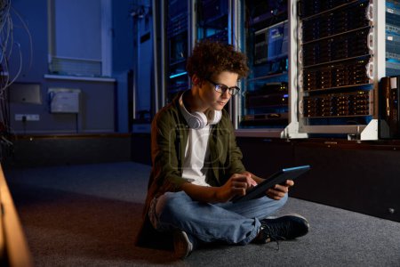 Photo for Concentrated teenager IT worker sitting on floor of modern server room working on tablet searching software virus - Royalty Free Image