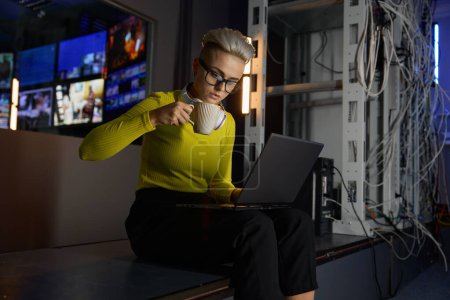 Téléchargez les photos : Jeune femme hipster travaillant avec un ordinateur portable dans une salle de serveur de datacenter sombre vérifiant la connexion réseau - en image libre de droit
