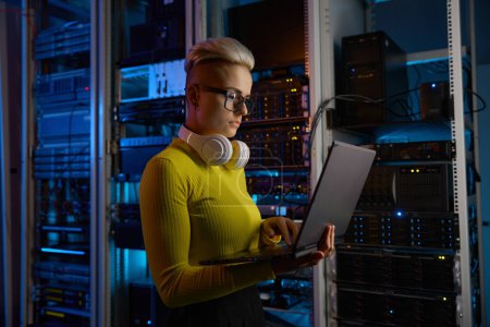 Side view of woman engineer working on laptop doing backups operation at network equipment in dark server room