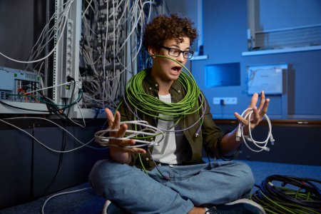 Photo for Shocked teenager geek wrapped in wire sitting on floor in data center. Problem with internet connection cables - Royalty Free Image