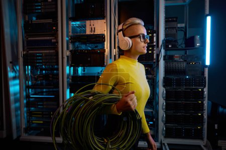 Téléchargez les photos : Jeune femme technicienne informatique portant des écouteurs portant faisceau de fils marchant à travers le rack du serveur dans le centre de Big Data à l'intérieur - en image libre de droit