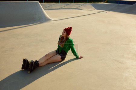Téléchargez les photos : Fatiguée mais satisfaite adolescente portant des patins à roulettes assis sur un rouleau d'asphalte. Après l'entraînement sportif actif - en image libre de droit