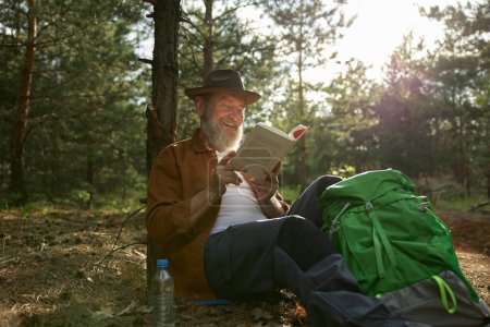 Photo for Happy senior man reading funny book during rest after hiking in forest. Travel, wellness and vacation on retirement - Royalty Free Image