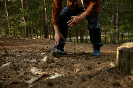 Téléchargez les photos : Homme âgé s'est foulé la jambe pendant une randonnée en plein air dans la forêt. Voyageur retraité souffrant de mal de pied ayant un problème de santé tout en marchant sentier sur la nature - en image libre de droit