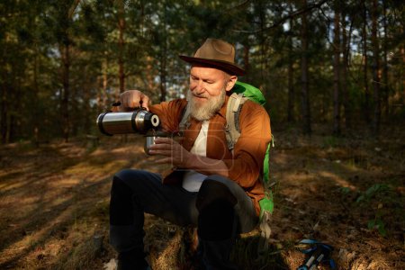 Foto de Senderista hombre mayor beber té caliente de la taza de termo durante el descanso en el bosque. Senderismo fuera en la jubilación - Imagen libre de derechos