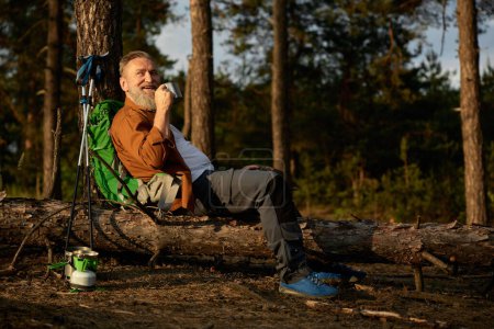 Téléchargez les photos : Randonneur senior prenant une pause café pendant l'aventure de marche active en plein air dans la forêt de pins. Un retraité boit une boisson chaude après le déjeuner. Week-ends sur la nature pendant la retraite - en image libre de droit