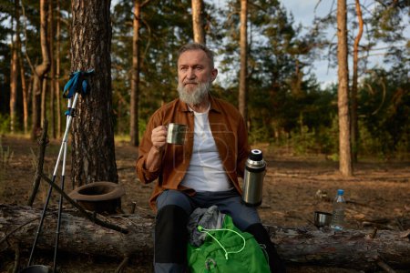 Foto de Hombre maduro sentado y bebiendo café caliente o té del termo mientras descansa en el bosque después de una aventura de senderismo - Imagen libre de derechos