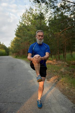 Photo for Senior man jogger stretching leg preparing for running. Flexible sportsman doing workout in park. Physical training outdoors - Royalty Free Image