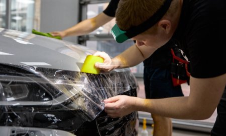 Polyurethane vinyl film pasting on car hood procedure closeup. Team of automotive master working at garage service