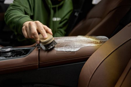 Téléchargez les photos : Nettoyage intérieur de voiture en cuir avec détergent mousse et brosse. Travailleur dans le lavage automatique des sièges d'auto à l'intérieur. Service de détail automatique - en image libre de droit