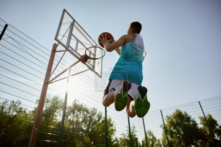 Photo for Teenager player throwing ball into basketball hoop, shot in motion, bottom view. Playing sport game on outdoor court - Royalty Free Image